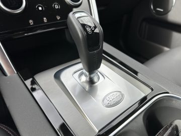 Close-up van de versnellingspook in een Land Rover Discovery Sport P300e, waarbij de omringende bedieningselementen en een deel van het gestroomlijnde dashboard aan de binnenkant worden benadrukt.