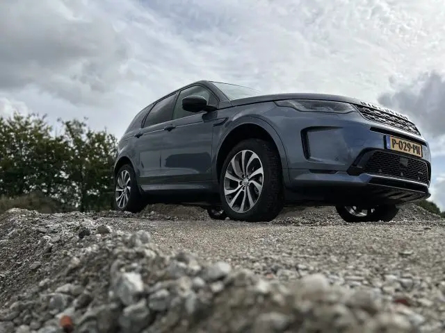 Een grijze Land Rover Discovery Sport P300e geparkeerd op een rotsachtig oppervlak met bomen en een bewolkte lucht op de achtergrond.