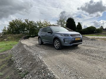 Op een grindheuvel staat een grijze Land Rover Discovery Sport P300e met Nederlands kenteken geparkeerd, omgeven door groen en een bewolkte lucht.