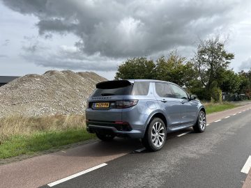 Een Land Rover Discovery Sport P300e staat op een bewolkte dag langs de kant van een verharde weg geparkeerd; op de achtergrond zijn een grote stapel grind en enkele bomen zichtbaar.