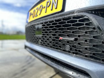 Close-up van de grille van een Land Rover Discovery Sport P300e met een gele kentekenplaat met daarop "029-PN" en een "R Dynamic"-badge, waterdruppels zichtbaar op het oppervlak, met een onscherpe achtergrond van gras en lucht.