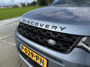 Close-up van de grille en kentekenplaat van een Land Rover Discovery Sport P300e, met waterdruppels op het oppervlak van de auto. Op het kenteken staat "P-029-PN.