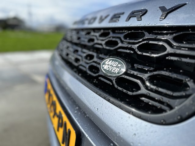 Close-up van een zilveren Land Rover Discovery Sport P300e-autogrille met zichtbare waterdruppels. Het merklogo en een deel van de kentekenplaat van het voertuig zijn zichtbaar. De achtergrond is wazig.