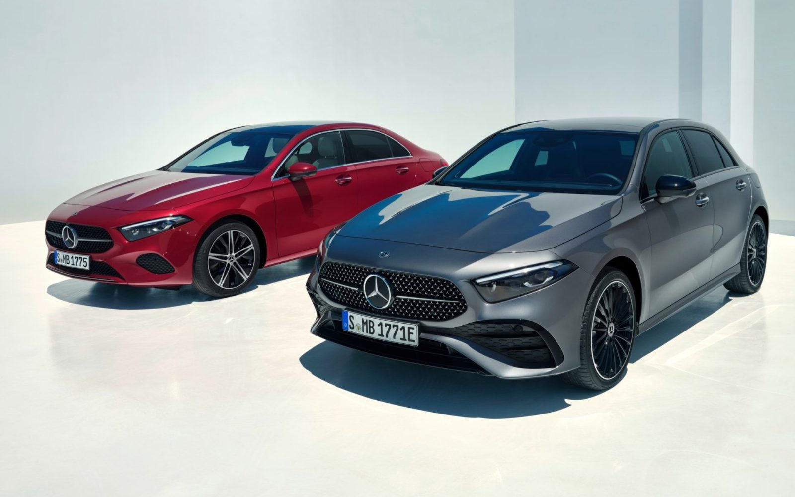 Twee Mercedes-Benz A-Klasse-auto's staan geparkeerd op een witte ondergrond. De ene is rood en de andere is grijs, beide vanuit een kleine hoek gezien vanaf de voorkant.