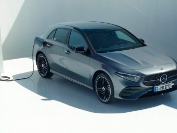 Een zilveren Mercedes-Benz A-Klasse elektrische auto staat geparkeerd en wordt opgeladen bij een wit laadstation in een minimalistische, lichtrijke ruimte.