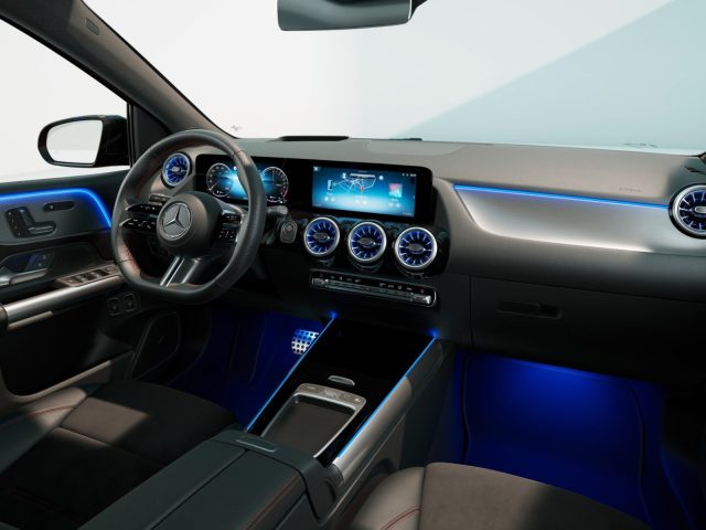 Modern auto-interieur van de Mercedes-Benz A-Klasse met een strak dashboard met digitaal display, blauwe sfeerverlichting en drie ronde ventilatieopeningen. Op het stuur prijkt trots het Mercedes-Benz-logo.