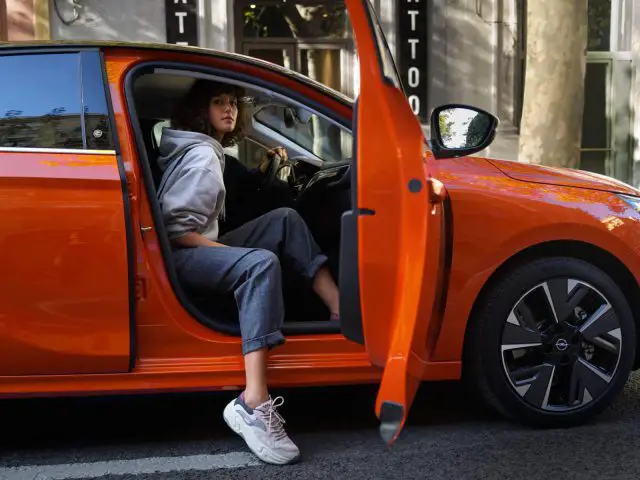 Een persoon in vrijetijdskleding zit op de bestuurdersstoel van een oranje Opel met de deur open, geparkeerd in een stadsstraat.