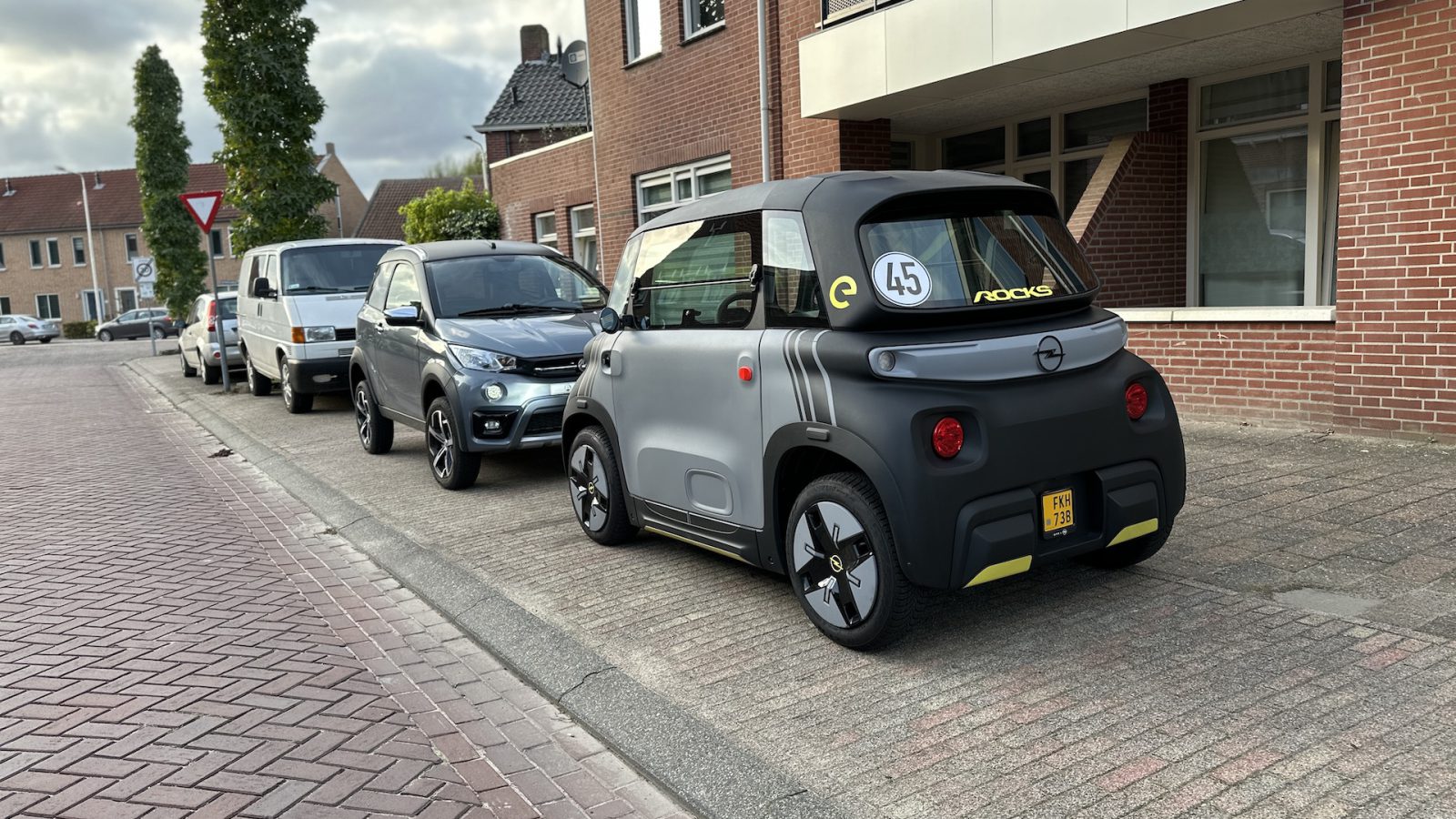 Een compacte Opel Rocks-e met het nummer 45 op de achterruit staat naast andere auto's geparkeerd in een woonstraat met bakstenen gebouwen.