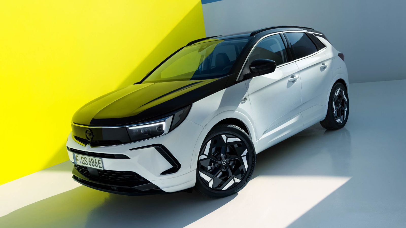 Een witte Opel Grandland GSe met zwarte accenten staat geparkeerd in een helder verlichte, moderne ruimte met gele en grijze muren. Het voertuig heeft een strak design en getinte ramen.