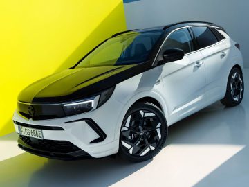 Een witte Opel Grandland GSe met zwarte accenten staat geparkeerd in een helder verlichte, moderne ruimte met gele en grijze muren. Het voertuig heeft een strak design en getinte ramen.