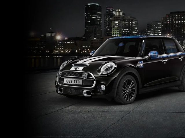 Een zwarte Mini Cooper, die zijn automatische personaliserende kenmerken laat zien, staat 's nachts geparkeerd op een betonnen oppervlak, met een verlichte skyline van de stad op de achtergrond.
