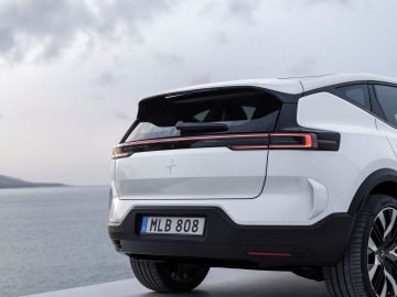 Close-up van de achterkant van een witte SUV, mogelijk een Polestar 3, met een opvallend achterlichtontwerp, geparkeerd nabij een watermassa. Het voertuig is voorzien van een Europees kenteken met de tekst "MLB 808.