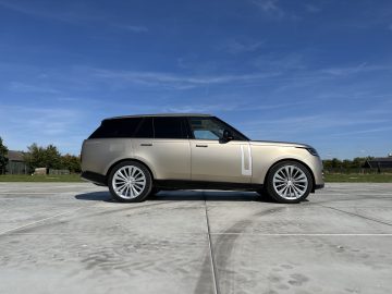 Een beige Range Rover staat geparkeerd op een betonnen ondergrond tegen een achtergrond van een helderblauwe lucht en verre bomen.