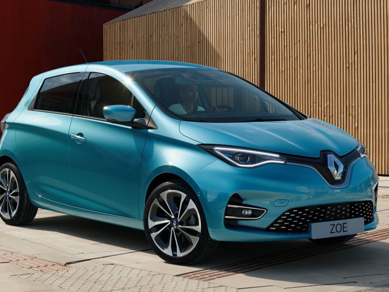 Een turquoise Renault Zoe geparkeerd op een verhard terrein voor charmante houten en bakstenen gebouwen, die naadloos opgaan in de schilderachtige omgeving.