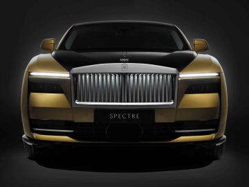 Een vooraanzicht van een goud-zwarte Rolls-Royce Spectre-luxeauto met een prominente grille en strakke koplampen. Op het kenteken staat 'Spectre.