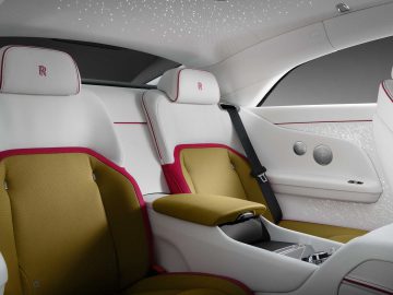 Interieur van een luxe Rolls-Royce Spectre met witte en olijfgroene stoelen, rode accenten, armleuning, veiligheidsgordel en sterrenplafondeffect. De hoofdsteunen zijn voorzien van een logo met een "R" erop geborduurd.