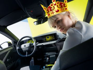 Persoon die een digitale kroon draagt in een moderne auto, naar achteren gericht terwijl hij op de bestuurdersstoel zit, waarschijnlijk onderdeel van een onderzoek naar rijgedrag. Heldergele achtergrond gezien door de ramen.