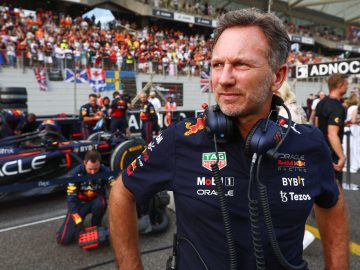 Een man, gekleed in een raceteamuniform en hoofdtelefoon, staat op een circuit naast de raceauto van Oscar Piastri, met mensen en een menigte op de achtergrond.