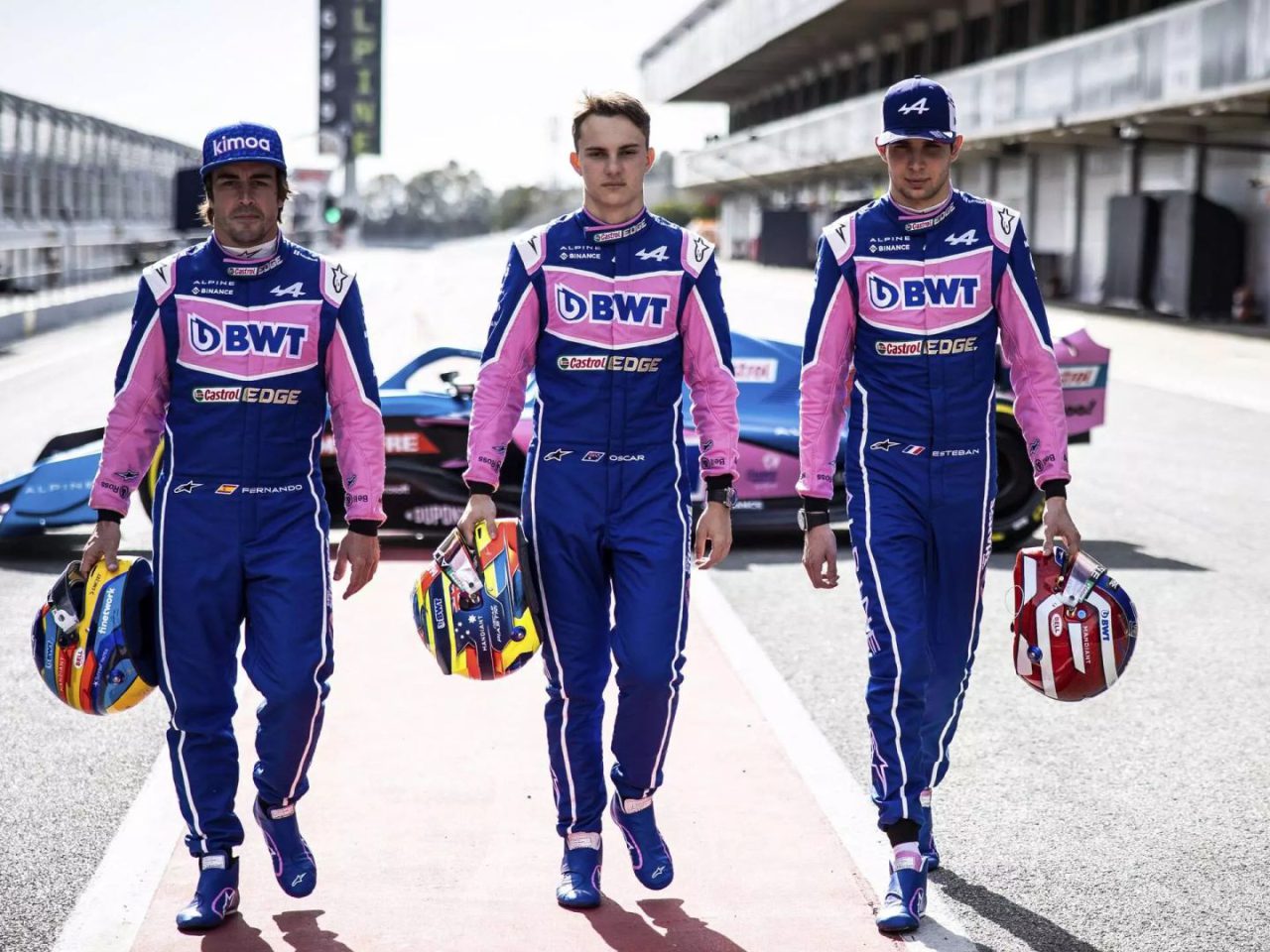 Drie autocoureurs in blauwe en roze uniformen, waaronder Oscar Piastri, lopen elk met een helm op een racebaan.