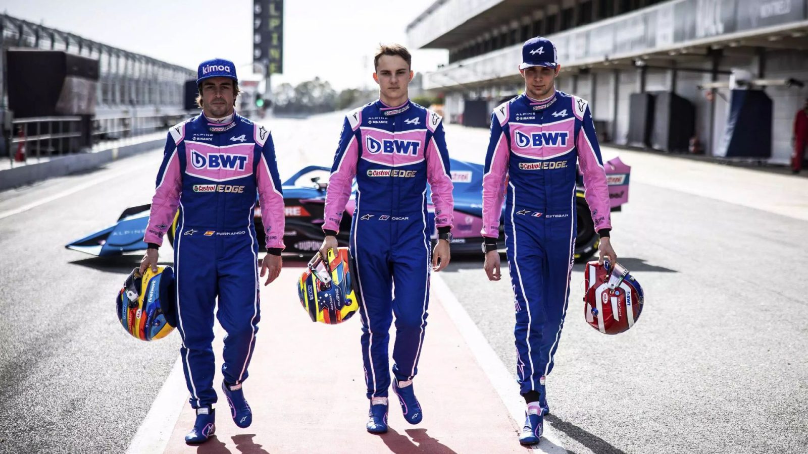 Drie autocoureurs in blauwe en roze uniformen, waaronder Oscar Piastri, lopen elk met een helm op een racebaan.
