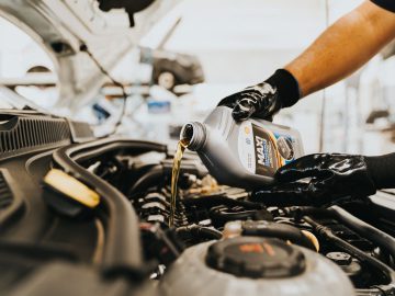 Een persoon die handschoenen draagt, giet motorolie uit een fles in een automotor in een autoserviceomgeving, wat het belang van goed onderhoud illustreert. Rijden zonder olievuldop: hoe erg is dat?