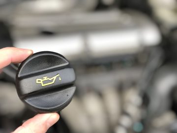 Een close-up van een hand die een oliedop vasthoudt met daarop een geel oliekan-symbool en de vraag stelt: "Rijden zonder olievuldop: hoe erg is dat?" De motor van de auto is zichtbaar op de onscherpe achtergrond.