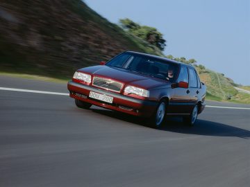 Een rode Volvo 850 vierdeurs sedan rijdt op een bochtige weg met een heuvelachtige achtergrond, vastgelegd in beweging. Dit is een vooraanzicht van het voertuig.