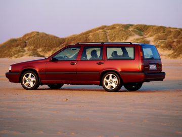 Een rode Volvo 850 stationwagen staat geparkeerd op een zandstrand met met gras begroeide zandduinen op de achtergrond.