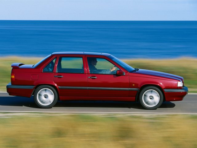 Een rode Volvo 850 sedan rijdt over een kustweg met de oceaan op de achtergrond.