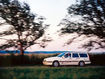 Een witte Volvo 850 stationwagen met het opschrift "Drive" rijdt over een landelijke weg met vage bomen en het platteland op de achtergrond.