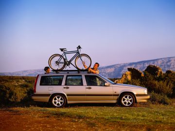 In een landelijk landschap staat een beige Volvo 850 stationwagen geparkeerd met een fiets op het imperiaal. Twee mensen zetten de fiets vast. Heuvels en struiken zijn zichtbaar op de achtergrond onder een heldere hemel.