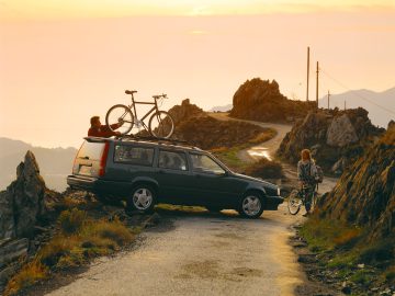 Op een smal bergweggetje staat bij zonsondergang een Volvo 850 met twee fietsen op de imperiaal geparkeerd. Twee mensen, één naast de auto en één met een fiets in de hand, zijn met elkaar in gesprek.