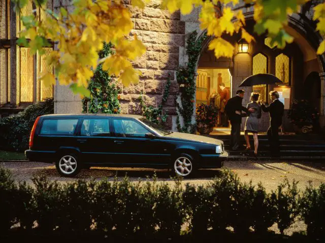 Een zwarte Volvo 850 stationwagen staat geparkeerd voor de ingang van een stenen gebouw, terwijl verschillende mensen onder een paraplu bij de deuropening staan. Het tafereel wordt omlijst door herfstgebladerte.