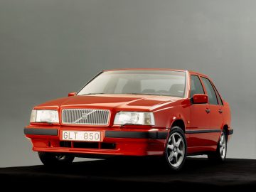 Tegen een grijze achtergrond wordt een rode Volvo 850 sedan met een kentekenplaat met de tekst "GLT 850" getoond.