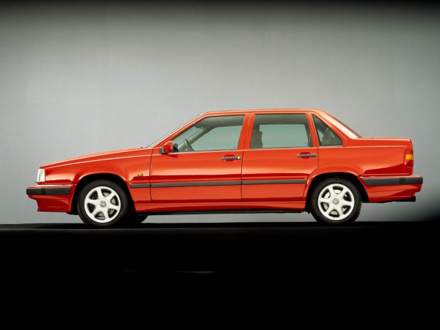 Zijaanzicht van een rode Volvo 850 sedan met een vierkant ontwerp, vier deuren en lichtmetalen velgen, tegen een effen achtergrond.