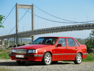 Een rode Volvo 850 sedan staat op een heldere dag buiten geparkeerd voor een grote hangbrug.