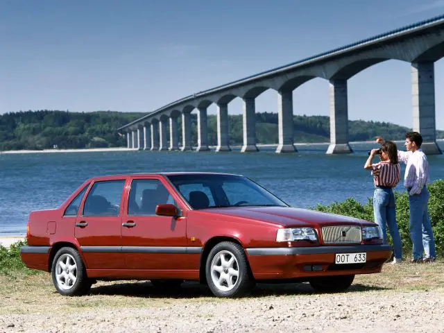 Een rode Volvo 850 sedan staat geparkeerd bij een watermassa met een brug op de achtergrond. Er staan twee mensen vlakbij, genietend van het landschap.