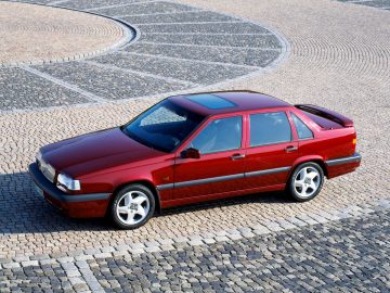 Een rode vierdeurs Volvo 850 sedan staat geparkeerd op een geplaveid oppervlak met een patroon. De auto heeft een schuifdak en lichtmetalen velgen.