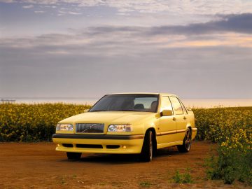 Een gele Volvo 850 sedan staat geparkeerd op een onverharde weg te midden van een veld met gele bloemen, met een bewolkte lucht op de achtergrond.