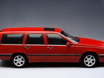 Een rode Volvo 850 stationwagen met vijf deuren, zilverkleurige lichtmetalen velgen en een schuifdak, vanaf de zijkant gezien tegen een neutrale achtergrond.