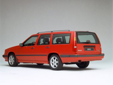 Een rode Volvo 850 stationwagen wordt tentoongesteld in een studioomgeving, waarbij het strakke zij- en achteraanzicht tegen een zuivere witte achtergrond wordt benadrukt.