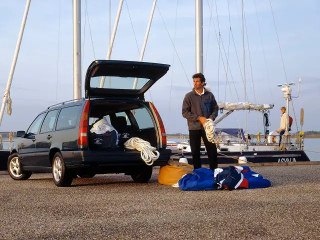 Een man staat naast een open kofferbak van een Volvo 850, gevuld met touwen en tassen in een jachthaven. Twee mensen zitten op een zeilboot die vlakbij is aangemeerd.