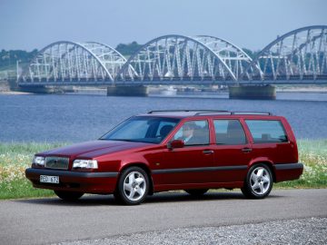 Een rode Volvo 850 stationwagen staat geparkeerd op een weg nabij een grote watermassa, met een gebogen stalen brug op de achtergrond.