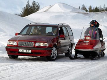 Een rode Volvo 850 stationwagen die over een besneeuwde weg rijdt naast een sneeuwscooter die wordt bestuurd door een persoon die een sneeuwhelm draagt.