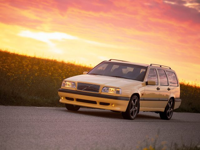 Een gele Volvo 850 stationwagen staat geparkeerd op een landelijke weg tijdens een kleurrijke zonsondergang.