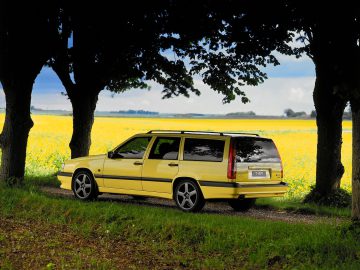 Op een landelijk pad tussen de bomen staat een gele Volvo 850 stationwagen geparkeerd, met op de achtergrond een veld met gele bloemen.