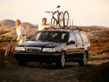 Een man bevestigt fietsen aan de imperiaal van een zwarte Volvo 850 stationwagen, terwijl een vrouw vlakbij staat met een andere fiets. De setting lijkt een rotsachtig buitenlandschap te zijn.