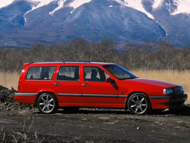 Een rode Volvo 850 stationwagen staat geparkeerd op een onverharde weg met een besneeuwde bergketen op de achtergrond.