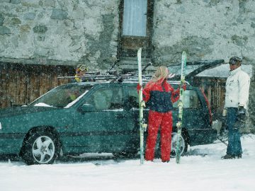 Een vrouw in rode skikleding en twee mannen leggen tijdens sneeuwweer hun skiuitrusting klaar naast een groene Volvo 850, met een stenen gebouw op de achtergrond.