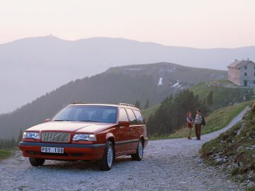 Een rode Volvo 850 stationwagen staat geparkeerd op een onverharde weg in een bergachtig gebied terwijl twee mensen vlakbij lopen; op de achtergrond is een huis zichtbaar.